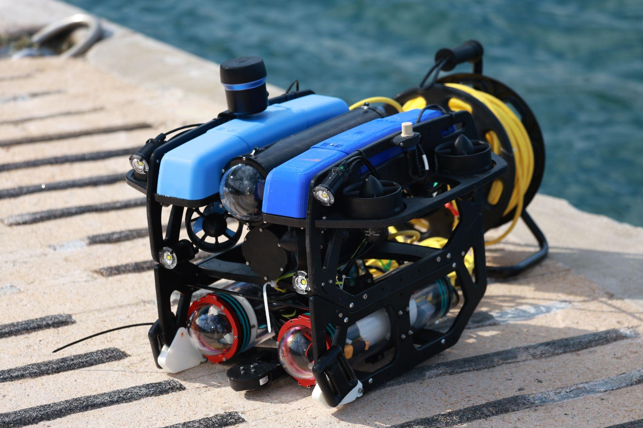 Blue multi-sensor underwater ROV with camera housings, scanning sonar and a tether reel beside the water